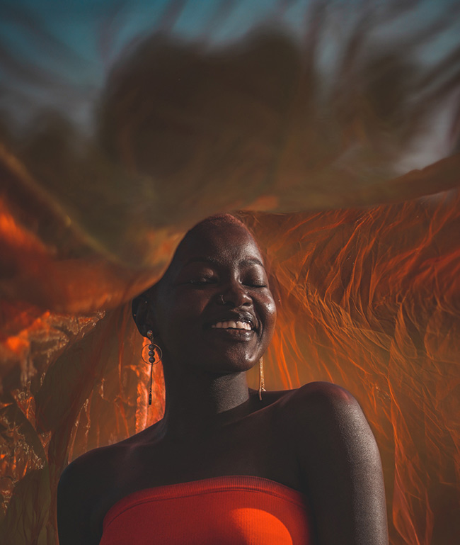 A person wearing a strapless red top smiles with their eyes closed. They are surrounded by flowing orange fabric that catches the light, creating a warm, artistic backdrop. The sky is visible in the background.