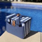 A blue and white striped cooler bag with brown leather accents sits on the edge of a swimming pool, with the water glistening in the sunlight.
