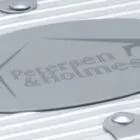 Close-up of a silver metal surface with an oval plaque featuring the text Petersen & Holmes. The metal has a ribbed texture and is fastened with bolts.