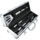 A silver metal case containing stainless steel barbecue utensils, including a spatula, tongs, and a fork, neatly secured with black straps. The case has a handle and latches.