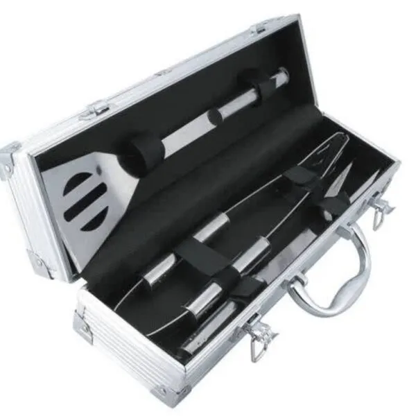 An open silver metal case containing barbecue tools on black padding. The set includes a spatula, tongs, and a fork, all with metallic handles. The case has a handle and latches for closing.
