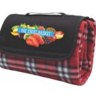 Folded red and black checkered picnic blanket with a black flap. The flap features colorful fruit graphics and the text The Fruit Basket.