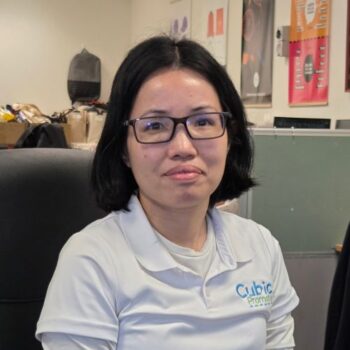 Meet the team: A woman with glasses sits in an office, wearing a white Cubic Promote shirt.