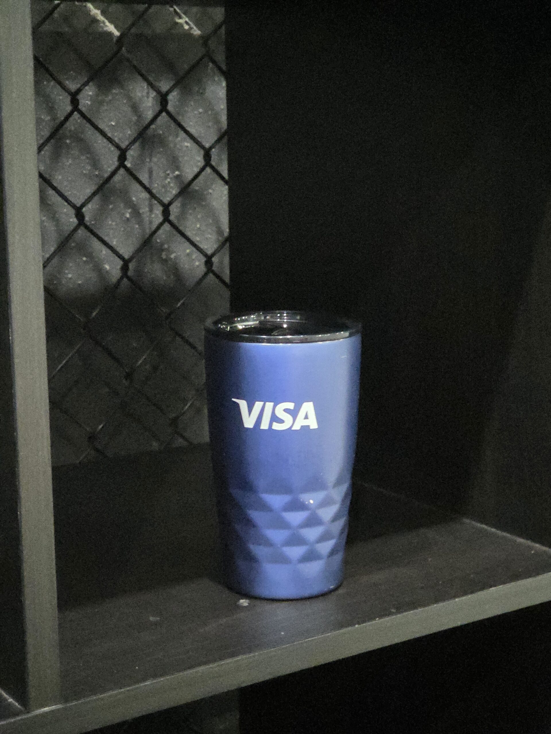 A blue tumbler with a VISA Australia logo sits on a dark shelf.
